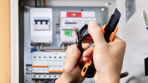 A pair of hands hold up a knife with some wires while pulling back the plastic cover. The person is working in front of a fuse box. 