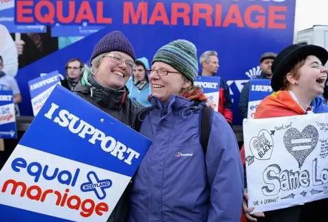 Getty Images A group of women close to camera in cold weather jackets and woolly hats holding banners saying "I support equal marriage". They are laughing and cheering. There are men with similar banners in the background.