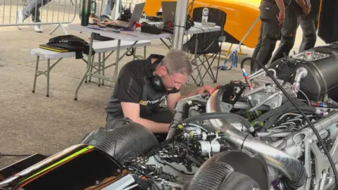 A photograph of an engineer kneeling beside the engine of the vehicle