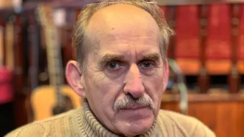 A man with a moustache wearing a brown jumper looks directly to the camera.