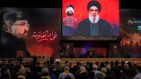 Reuters Hezbollah supporters listen to a speech by the group's leader Hassan Nasrallah (right), at a memorial ceremony for senior commander Fuad Shukr (left), after the latter was killed in an Israeli strike (6 August 2024)