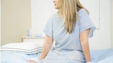 A woman wearing a white and blue hospital gown sits on the edge of a patient bed with her legs hanging down and her hands resting on the edge. She is looking to the left. The image is cut off above her nose. She has long, straight blonde hair.