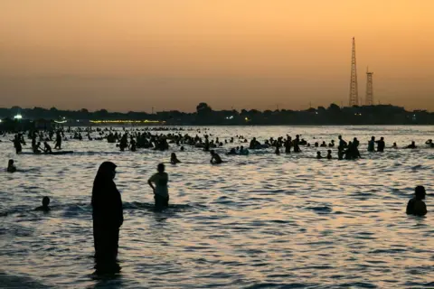 MAHMUD TURKIA / AFP / GETTY IMAGES People swim in the sea at sunset. A woman wearing an abaya and hijab can be seen in the foreground standing in the water up to her ankles.