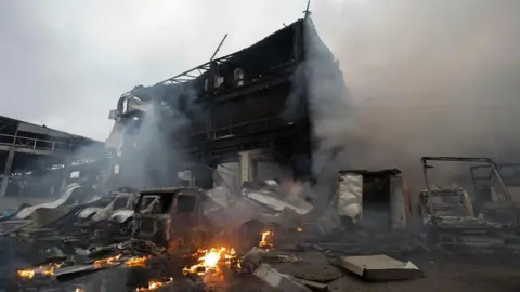 EPA Flames are seen next to burnt out vehicles and a bombed building. Grey smoke billows into a a cloudy sky.