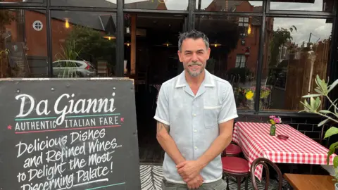 BBC/Richard Madden A man with a blue shirt, grey hair and goatee beard stands in front of a restaurant with large windows. A table has been covered with a red and white checked cloth and a large sign reads "Da Gianni - Authentic Italian Fare".