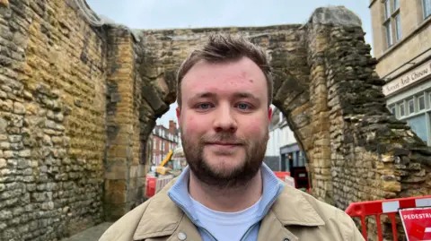 Councillor Joshua Wells is standing in front of a bricked Roman archway. The monument has mottled brickwork with green moss covered patches. Behind Joshua, work machinery can be seen in the background, including a JCB digger. Joshua is wearing a light beige coat with a light blue half zip jumper underneath. Joshua has blue eyes, a beard and brown hair. He is smiling. 
