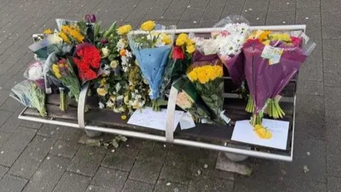 Bouquets of flowers cover a bench, along with cards and messages.