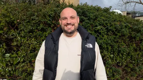 BBC Pete Fealey wearing a white jumper and a black gilet standing smiling in front of a green hedge. 