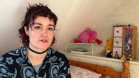 Niamh Tickner, a woman in her twenties with short, curly purple hair. She is sitting on the bed in her childhood bedroom, which has pink floral sheets and a pine headboard. Behind her are stuffed toys, a book and some trinkets. She has an eyebrow piercing and a piercing in her nose. She is wearing a dark blue paisley top and a black choker.