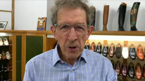 A man in a blue shirt stands in front of a large shoe rack with different colour oxford and brogue shoes on display. 