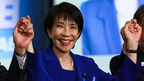 Getty Images Sanae Takaichi grins as she wears a blue dress with both her hands raised