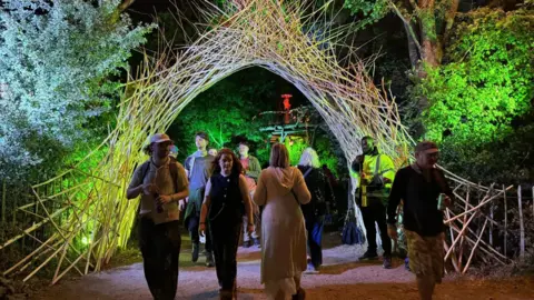 A wicker arch spans across a walkway in a wooded area. It is night-time and green lights are shining on the arch and trees. About a dozen people walk along the path.