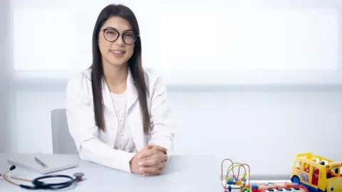 Courtesy of Diana Ramos / Photo by David Jiménez A woman with long brown hair wears a white medical coat and dark-framed glasses. She is seated indoors against a bright, predominantly white background that resembles a clinical setting. The image is a medium close-up portrait with soft lighting; no visible text, medical equipment, or other notable objects appear in the frame.