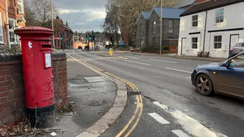 A red postbox is seen to the left of a suburban single-carriageway road, which is lined by houses and some trees, with traffic lights and a pedestrian crossing in the centre.