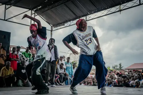 JOSPIN MWISHA / GETTY IMAGES People dance on stage wearing basketball jerseys and baggy trousers.
