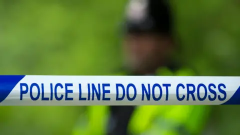 BBC A generic image of a blue-and-white police tape cordon, with an officer in a yellow high vis jacked pictured blurred in the background.
