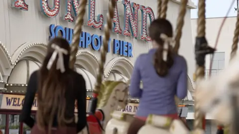 Two girls with their backs to the camera are sat on horses on a funfair carousel. They are both wearing cream hair bows looking at the front of the Southport pier sign