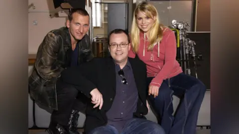 Christopher Eccleston is on the left of the photo, looking at the camera, wearing  a black leather jacket. Russell T Davies is in the centre, smiling at the camera, he is wearing glasses and has a black jacket, with a dark blue shirt. Billie Piper is on the right of the photo and is wearing a pink hoody and blue jeans. 