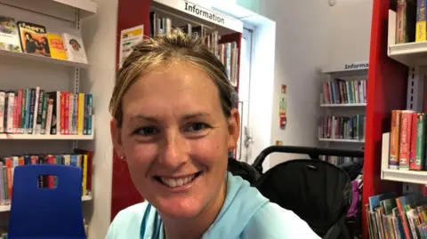 Vikki Irwin/BBC A woman with blonde hair is sitting in a library with a pushchair in the background