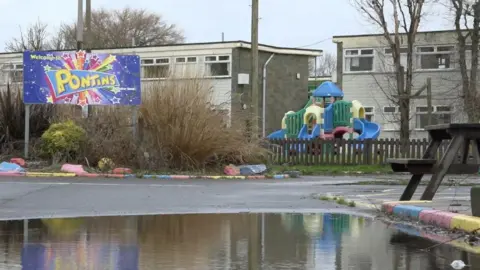 A picture from inside the holiday park, showing old blocks of flats and plastic play equipment that has faded primary colours. A large, red, yello and blue Pontins sign is on the left, and a large puddle of water is in the foreground.