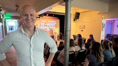 MP Max Wilkinson stood with his arms on his hips. There is a crowd of people watching a monologue to the right of him. He is smiling into the camera.