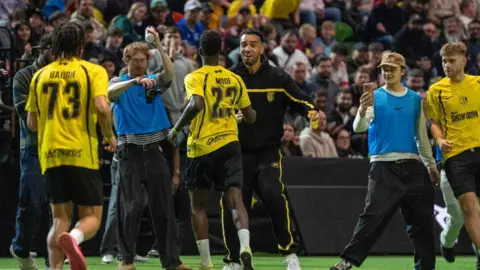 Baller League YouTube star Niko wears a black and yellow tracksuit while encouraging three members of his team on the football pitch. There are two photographers taking pictures of the group and a crowd of fans can be seen in the background 