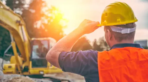 A stock photo of a construction site. A worker in a high-visibility vest is looking away from the camera, and his arm is bent up, touching the brim of his hard hat. A blurred digger is in the background, and the sun is shining in the background. 