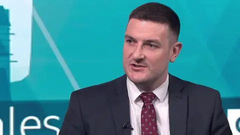 A man with short brown hair and a navy suit and red tie speaks on camera.