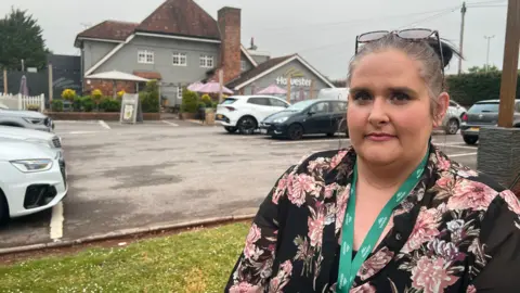 Helen Bill looks at the camera - she has a green lanyard on and wears a floral blouse. her hair is tied up in a bun. She stands in front of the Harvester car park and pub.