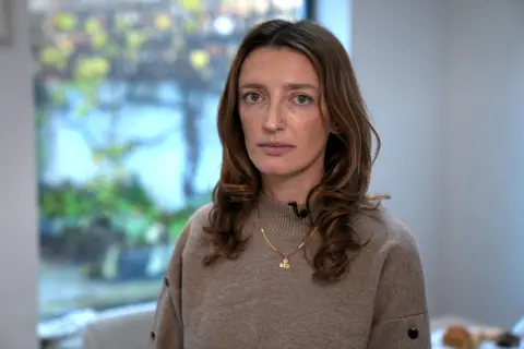 ಬಿಬಿಸಿ ಕ್ಲೇರ್ ಕಾರ್ಕೆರಿ, ಉದ್ದನೆಯ ಕೂದಲಿನ ಮಹಿಳೆ, ಬೀಜ್ ಜಂಪರ್ ಮತ್ತು ಚಿನ್ನದ ನೆಕ್ಲೇಸ್ ಧರಿಸಿ, ತನ್ನ ಊಟದ ಕೋಣೆಯಲ್ಲಿ ನಿಂತಿದ್ದಾಳೆ.