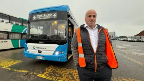 A man wearing an orange high visibility jacket and a blue coat underneath stands to the side of a blue and white liveried bus.