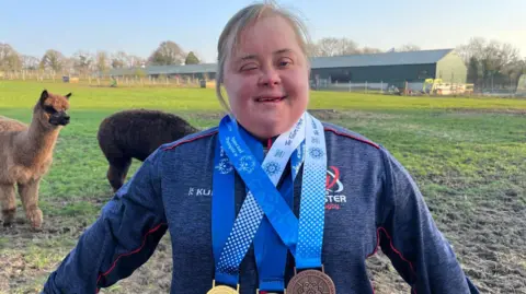 Lucy Best smiling at camera. She is wearing a grey jumper and has 3 medals around her neck. Two gold medals and a bronze. She is standing in a field.