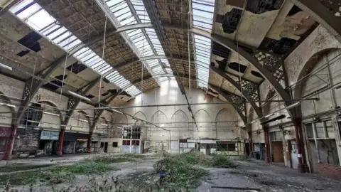 The inside of a disused indoor market building. Greenery is growing out of the grown and there is damage to the ceiling.