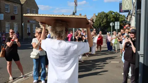 Sophie Tea Art A blonde woman in a white t-shirt carrying a mirror on her head as she walks down the pavement with about 20 people watching and taking photos on their phones.