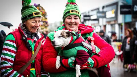 The Valley Evesham Two women standing side by side dressed in red, white and green elves costumes and green hats. The woman on the right is holding a white dog which has light brown ears which is also dressed in a red and green elf costume. 