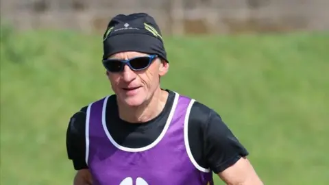 A man wearing a black hat and T-shirt, sunglasses and a purple bib, running with grassland in the background.
