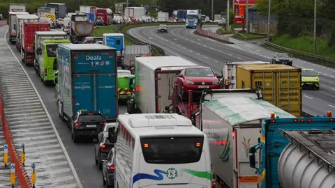 A very busy main road. There is a traffic jam on the road going in the direction away from the camera. On the side of the road coming toward the camera, traffic is clear. In the distance there is a sign advertising petrol prices. 