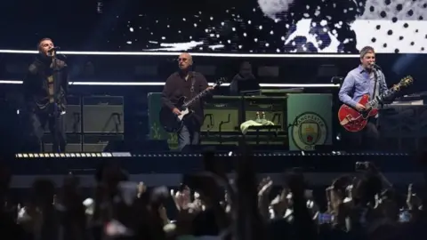 PA Media Liam and Noel Gallagher are singing into microphones as Oasis takes to the stage at the Principality Stadium, Cardiff, for the band's long-awaited reunion tour kicks off in Wales. Noel is also holding a guitar and a crowd of fans are in front of them.