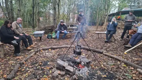 Two women and five men sat or stood in a circle in a wooded clearing, around a small wood fire with a metal frame hanging over it