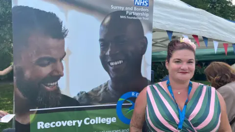 Emily Coady-Stemp/BBC The Recovery College's service manager, Nina Clayton Broom, pictured in front of a poster advertising the service. Nina is wearing a lanyard and a green/pink dress. Two men are seen smiling on the poster. 