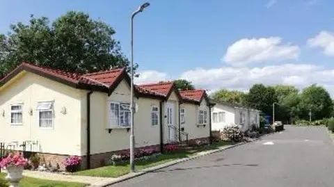 Alan Baldwin A row of single-storey park homes lines a quiet residential road at Penton Park, with cream-coloured homes, small front gardens and a streetlamp under a blue sky with scattered clouds.