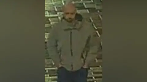 Merseyside Police A man is wearing a grey jacket and blue jeans and is walking along a street at night.