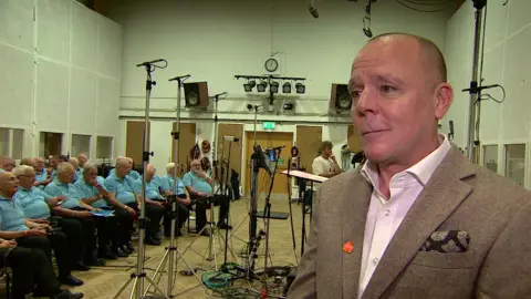 James Stevens is pictured in front of the choir. He is wearing a light brown suit jacket with pink open-necked shirt. He has a cravat in his breast picket and a red lapel badge.