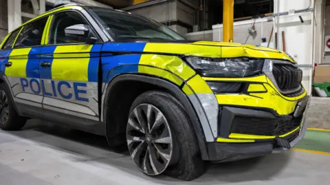 PSNI/PA Wire A damaged police vehicle - the vehicle is badly dented at the front and the tyre has come off the wheel. There are police markings on it which are blue and yellow with 'POLICE' written in blue on the side.