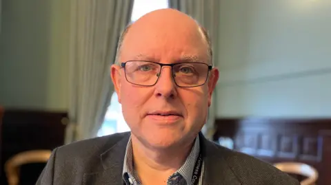 Councillor Andrew Jarvis. He wears glasses and has a bald head with grey hair at the sides. He is standing in what appears to be a council chamber, although the background is blurred.