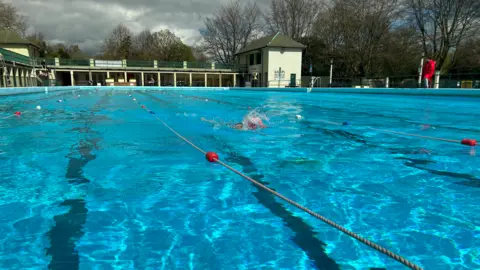 Emma Baugh/BBC Peterborough Lido, with clear blue water and a swimmer splashing while moving along a lane in the pool