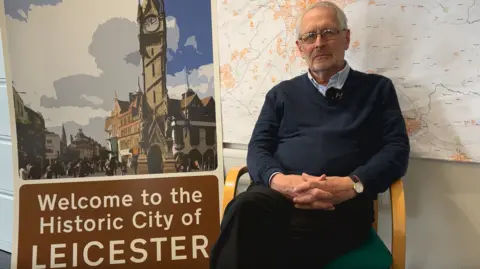 BBC A man in a chair sitting next to a brown "welcome to Leicester" road sign