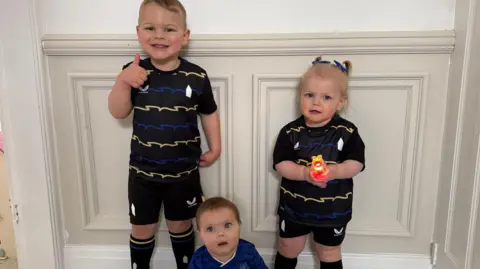PA A baby sits on the floor in front of two older children, a girl and a boy. All three are wearing football kits. 