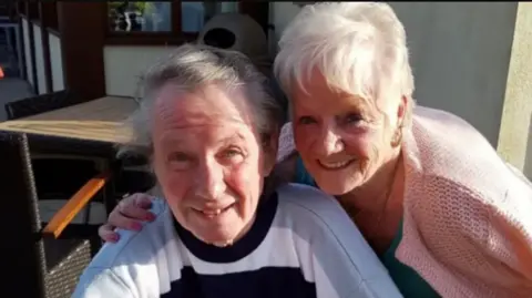 Merseyside Police handout Eric Greener, who has grey hair swept to the side and is wearing a blue and white striped sweatshirt, smiles at the camera while sitting in a chair outside a pub or restaurant. Sheila Jackson, who has short white hair and is wearing pink cardigan leans into the frame and smiles at the camera with her arm around Greener's shoulder.