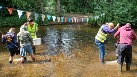 Two adult volunteers in high vis vests help children to do pond dipping in the shallow river. The children are seen from behind wearing wellies and shorts. There is bunting strung across the river.
